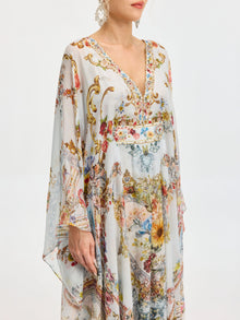 A woman models the Baroque Floral Print Silk Caftan Dress by Social Queen, featuring a flowing caftan silhouette, V-neck, wide sleeves, and colorful floral and baroque-inspired patterns against a plain backdrop.