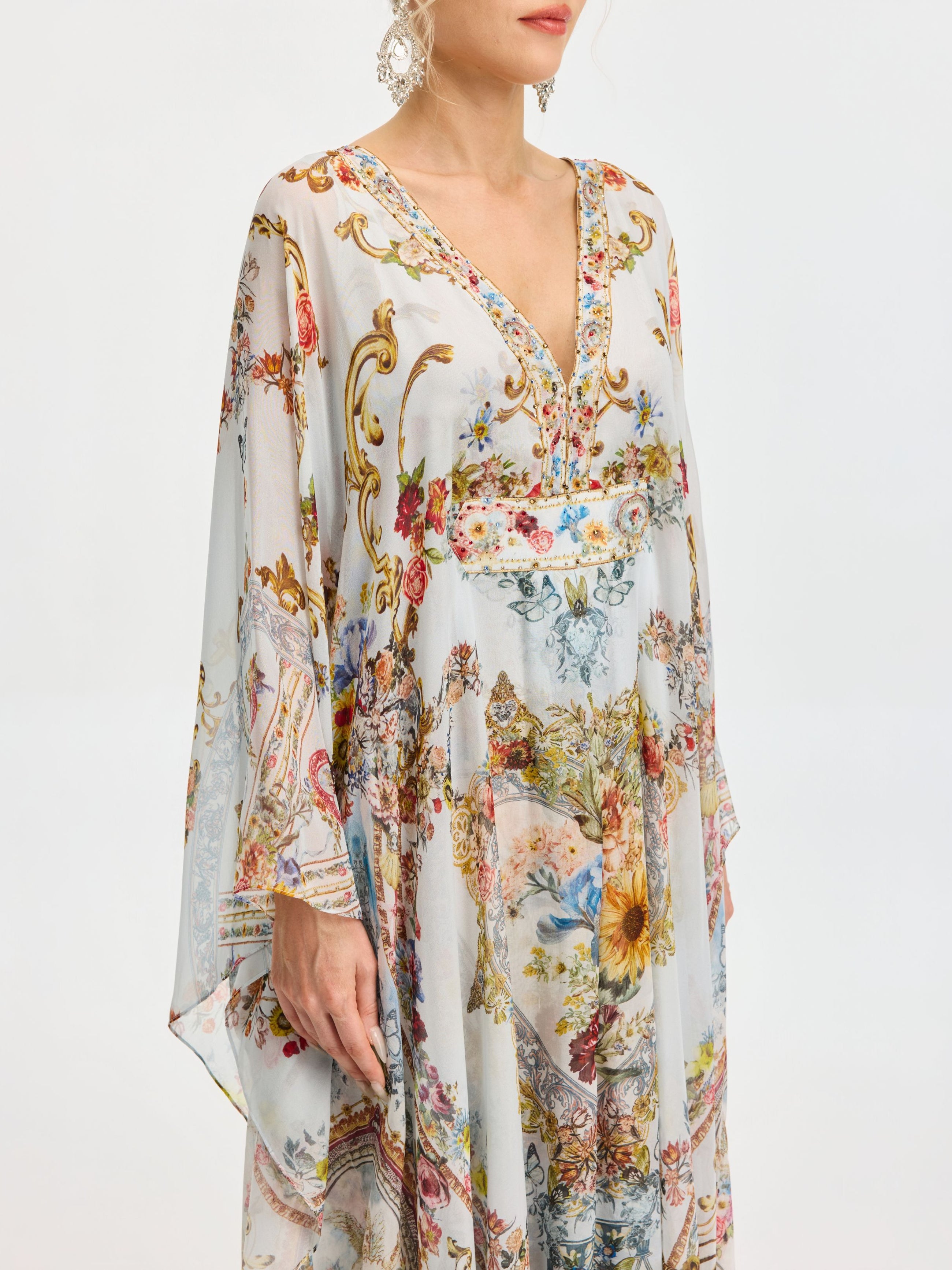 A woman models the Baroque Floral Print Silk Caftan Dress by Social Queen, featuring a flowing caftan silhouette, V-neck, wide sleeves, and colorful floral and baroque-inspired patterns against a plain backdrop.