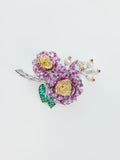 The Social Queen Camellia Bouquet Gemstone Brooch is a jeweled accessory shaped like flowers, adorned with pink and yellow gemstones, green gem leaves, and delicate pearl accents on a silver-colored base against a white background.