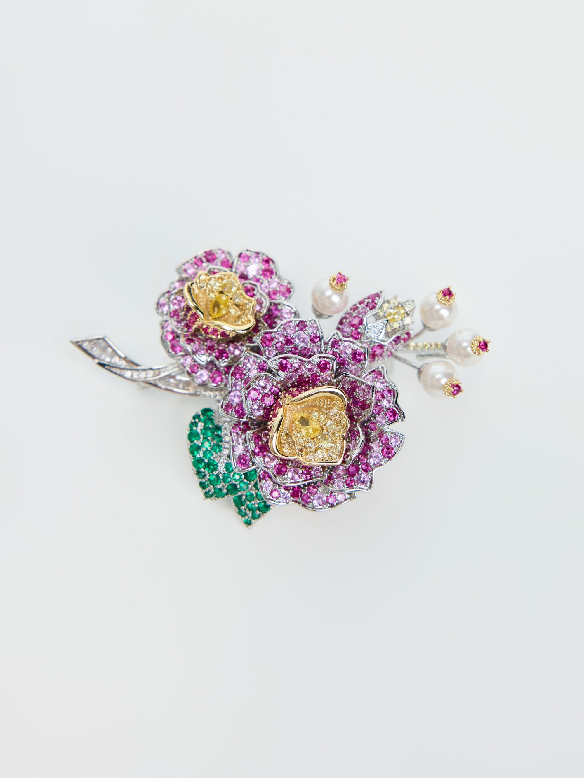 The Social Queen Camellia Bouquet Gemstone Brooch is a jeweled accessory shaped like flowers, adorned with pink and yellow gemstones, green gem leaves, and delicate pearl accents on a silver-colored base against a white background.