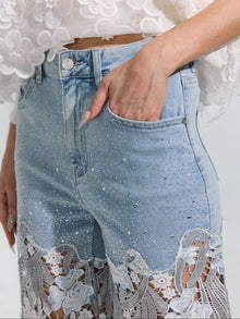 A person models the Social Queen Floral Lace Patchwork Pants—light blue stretch denim shorts adorned with rhinestone embellishments and finished with a white lace hem. One hand rests in the pocket. The top displays an intricate floral lace patchwork design.