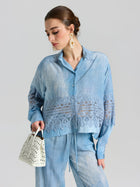 Woman wears the Social Queen Washed Silk Lace Detail Shirt in light blue, paired with relaxed fit pants and holding a white cut-out handbag, looking down against a plain background.