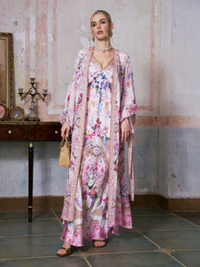 A woman stands indoors on a tiled floor, holding a small tan handbag and wearing the Social Queen Baroque Print Mulberry Silk Robe with Scarf over a long, floral-patterned pink and white dress, with a decorative table and candelabra in the background.