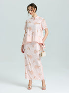 A woman wears the Social Queen Dragonfly Beaded Lace Midi Skirt, featuring light pink floral lace and beaded embroidery, paired with a matching top and holding a small purse against a plain background.