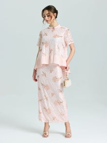 A woman wears the Social Queen Dragonfly Beaded Lace Midi Skirt, featuring light pink floral lace and beaded embroidery, paired with a matching top and holding a small purse against a plain background.