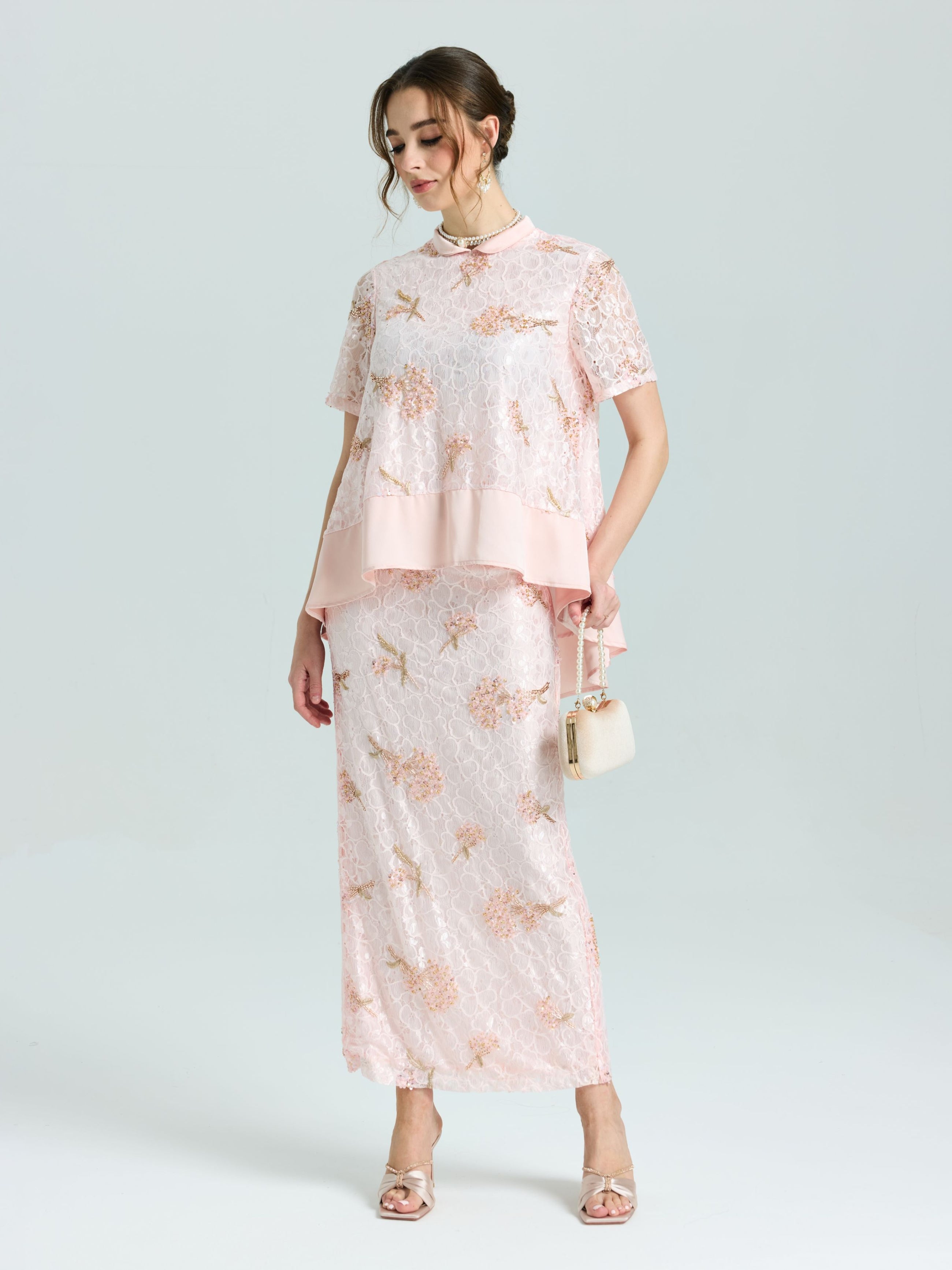 A woman wears the Social Queen Dragonfly Beaded Lace Midi Skirt, featuring light pink floral lace and beaded embroidery, paired with a matching top and holding a small purse against a plain background.