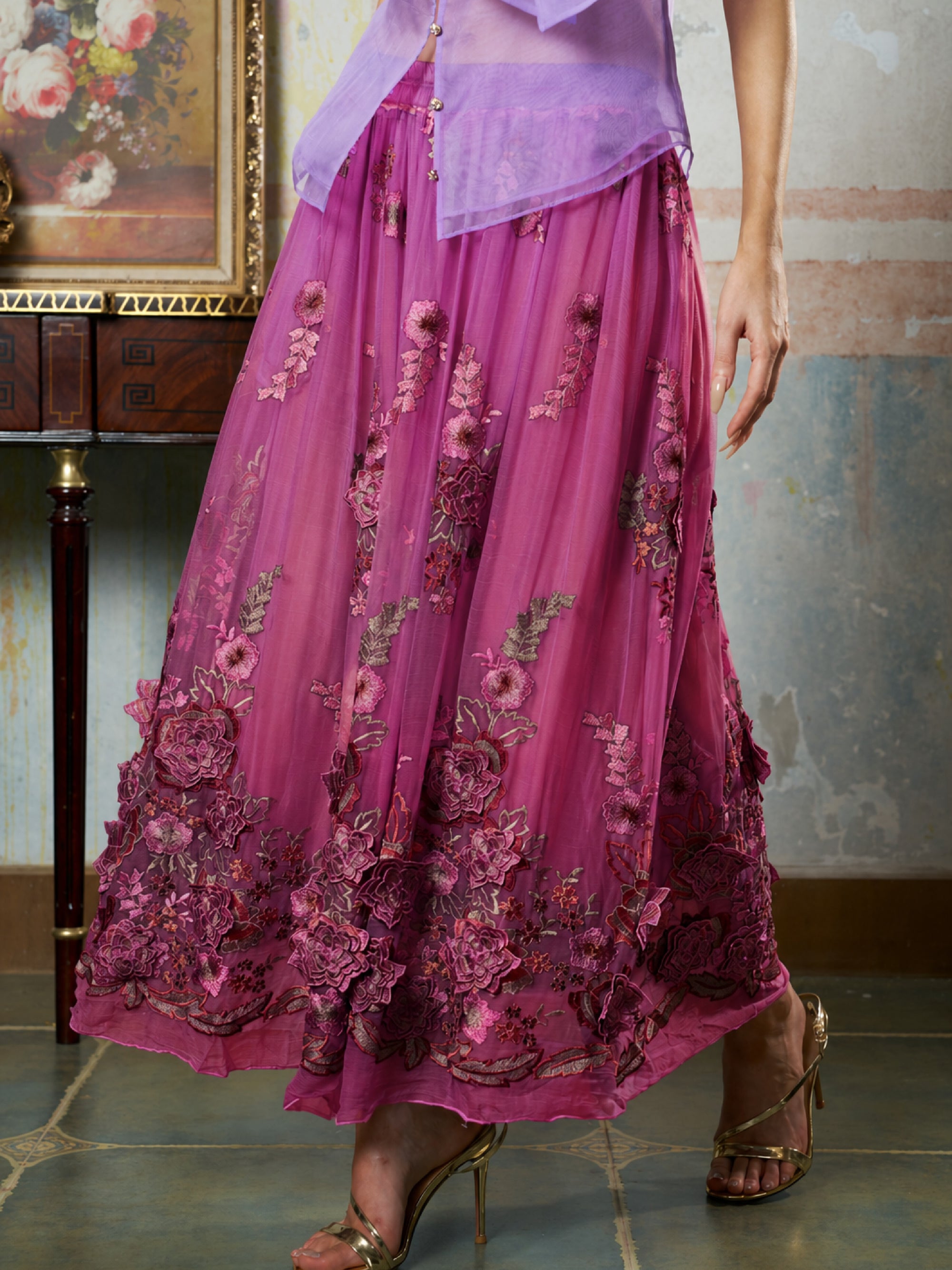 A person stands indoors by a wooden table and floral painting, wearing Social Queen’s 3D Floral Embroidered Silk Skirt in magenta with gold heeled sandals.