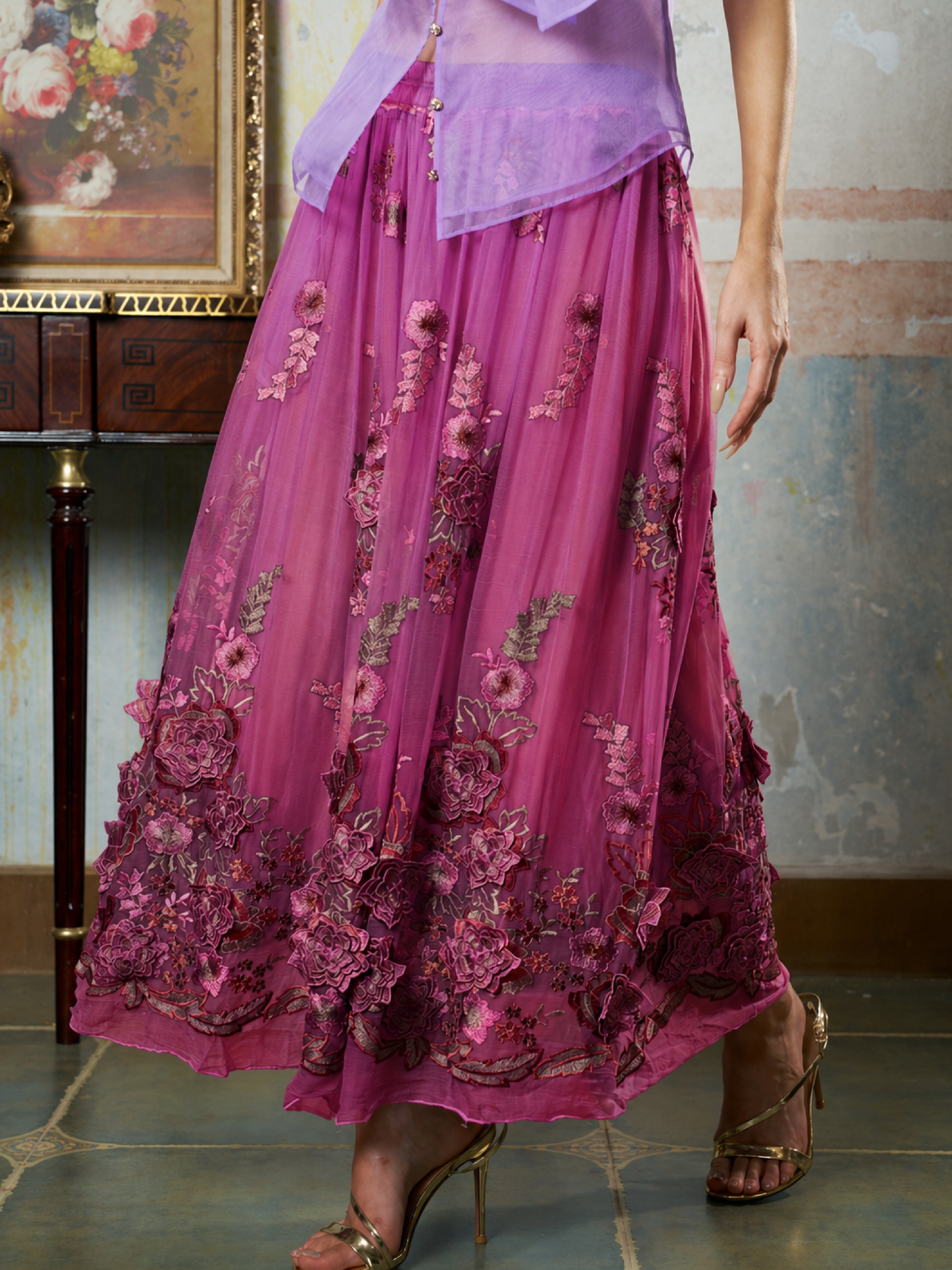 A person stands indoors by a wooden table and floral painting, wearing Social Queen’s 3D Floral Embroidered Silk Skirt in magenta with gold heeled sandals.