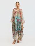 A woman models the Social Queen Floral Bird Silk Caftan Dress, featuring a V-neckline and sheer sleeves, styled with gold sandals and statement earrings—an elegant resort wear look against a plain background.