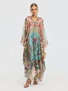 A woman models the Social Queen Floral Bird Silk Caftan Dress, featuring a V-neckline and sheer sleeves, styled with gold sandals and statement earrings—an elegant resort wear look against a plain background.
