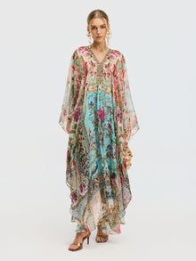 A woman models the Social Queen Floral Bird Silk Caftan Dress, featuring a V-neckline and sheer sleeves, styled with gold sandals and statement earrings—an elegant resort wear look against a plain background.