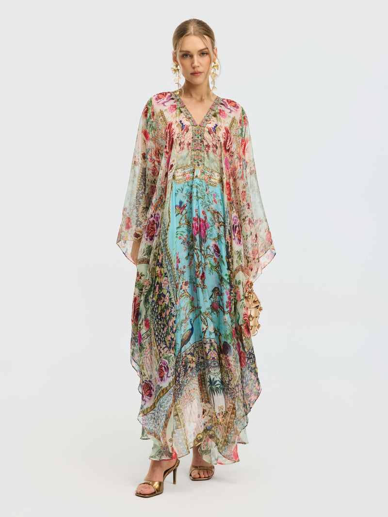 A woman models the Social Queen Floral Bird Silk Caftan Dress, featuring a V-neckline and sheer sleeves, styled with gold sandals and statement earrings—an elegant resort wear look against a plain background.