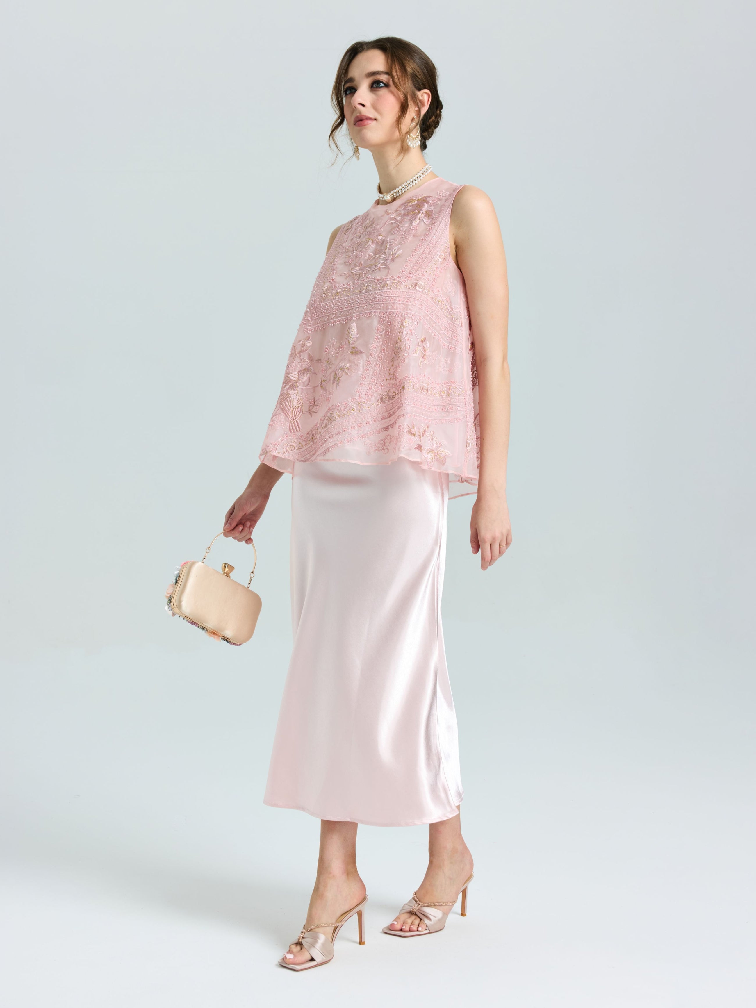 A woman models the Social Queen Satin A-Line Midi Skirt, paired with a sleeveless pink lace top, beige clutch, and high-heeled sandals, set against a plain light background.
