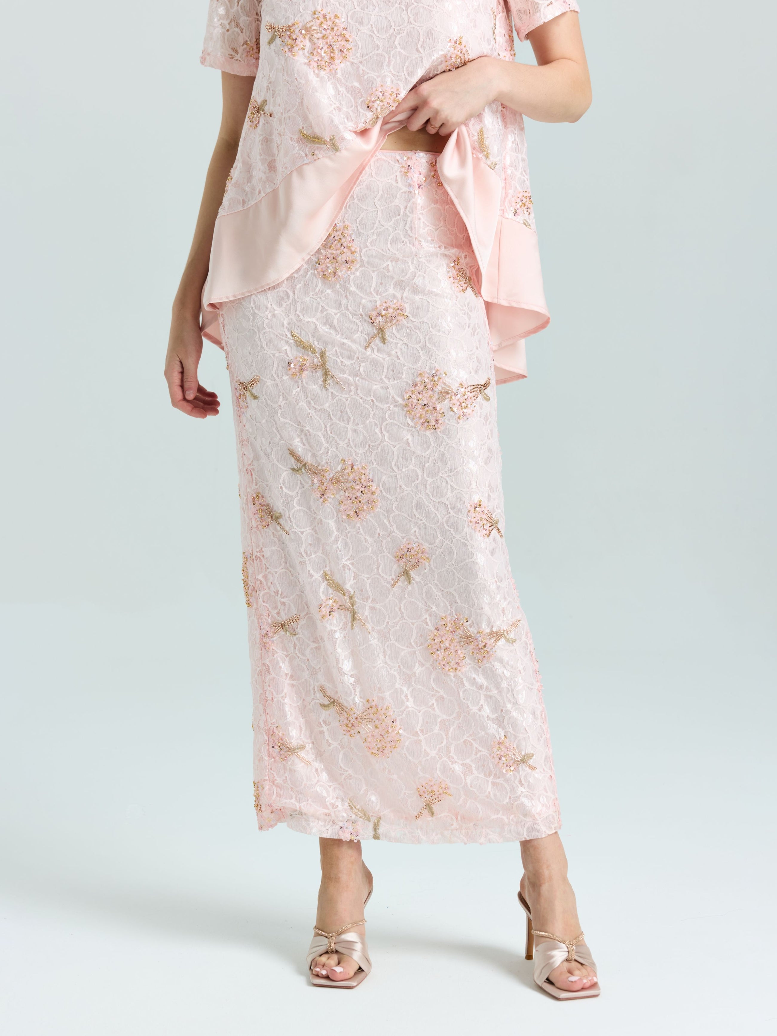 Only the lower half is visible: a person wears Social Queen's Dragonfly Beaded Lace Midi Skirt in light pink with beaded embroidery, styled in a straight silhouette, paired with open-toe heeled sandals and a matching top.
