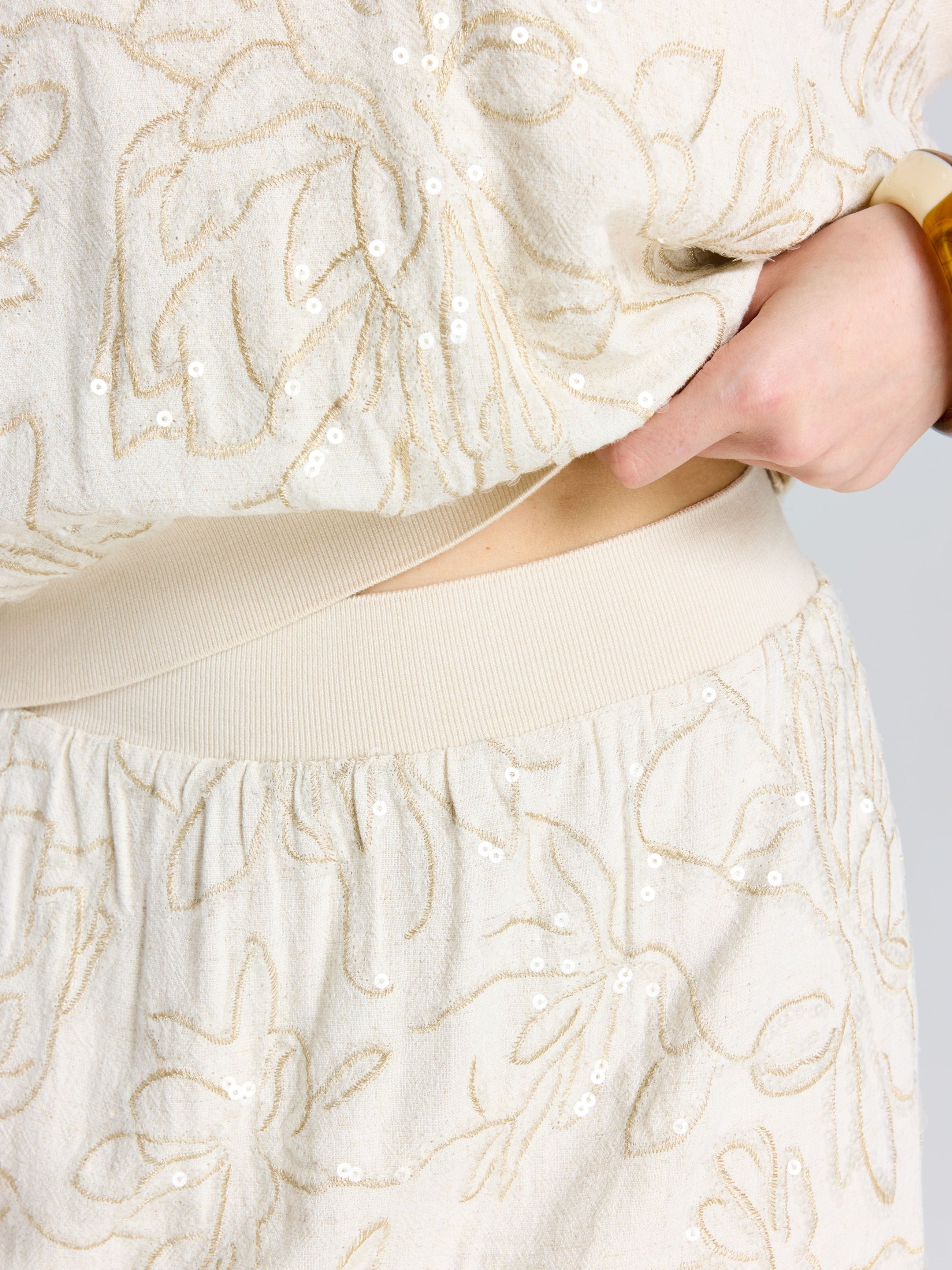 A person lifts the hem of the Social Queen Floral Embroidered Linen Midi Skirt in cream, revealing a hint of their bare waist.