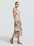 A woman wears the Social Queen Floral Embroidered Bow-Tie Dress, a light-colored A-line midi with short sleeves, paired with sandals and a small purse, standing against a plain background.
