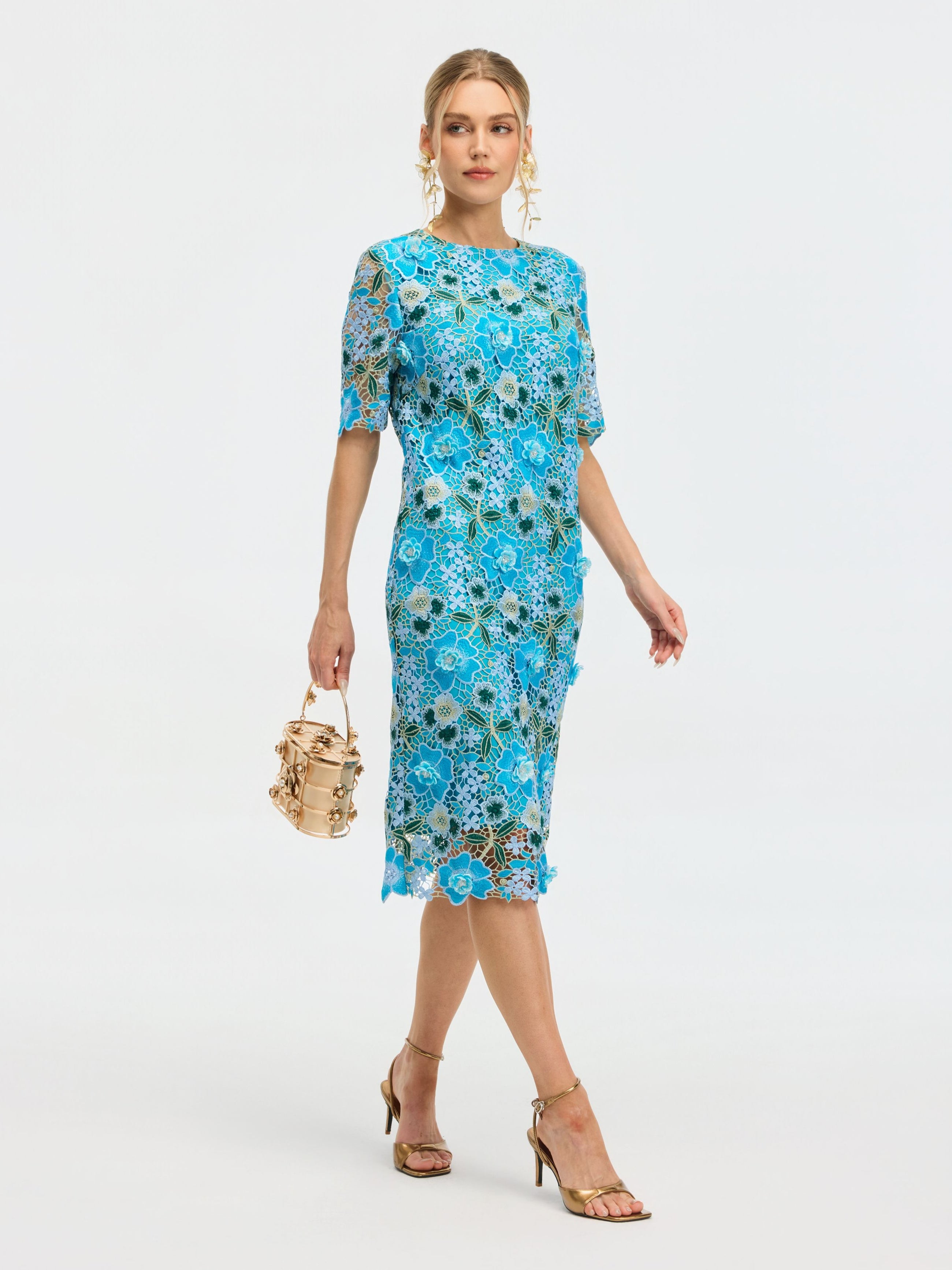 A woman wears the Social Queen Floral Appliqué Lace Shift Dress with gold earrings, gold heeled sandals, and carries a small beige handbag while walking against a plain white background.