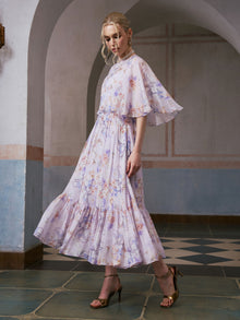 A woman wearing the Social Queen “Lavender Print Silk Dress” with flutter sleeves and a ruffled hem stands in a tiled corridor with arched walls, paired with gold high-heeled sandals.