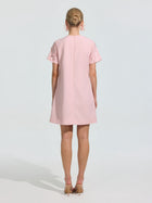 A woman faces away, wearing the Social Queen Beaded Floral Appliqué Dress—a short, light pink shift with short sleeves—paired with beige heels against a plain light background.