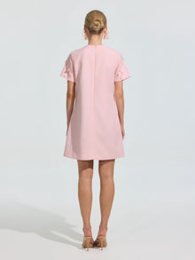 A woman faces away, wearing the Social Queen Beaded Floral Appliqué Dress—a short, light pink shift with short sleeves—paired with beige heels against a plain light background.