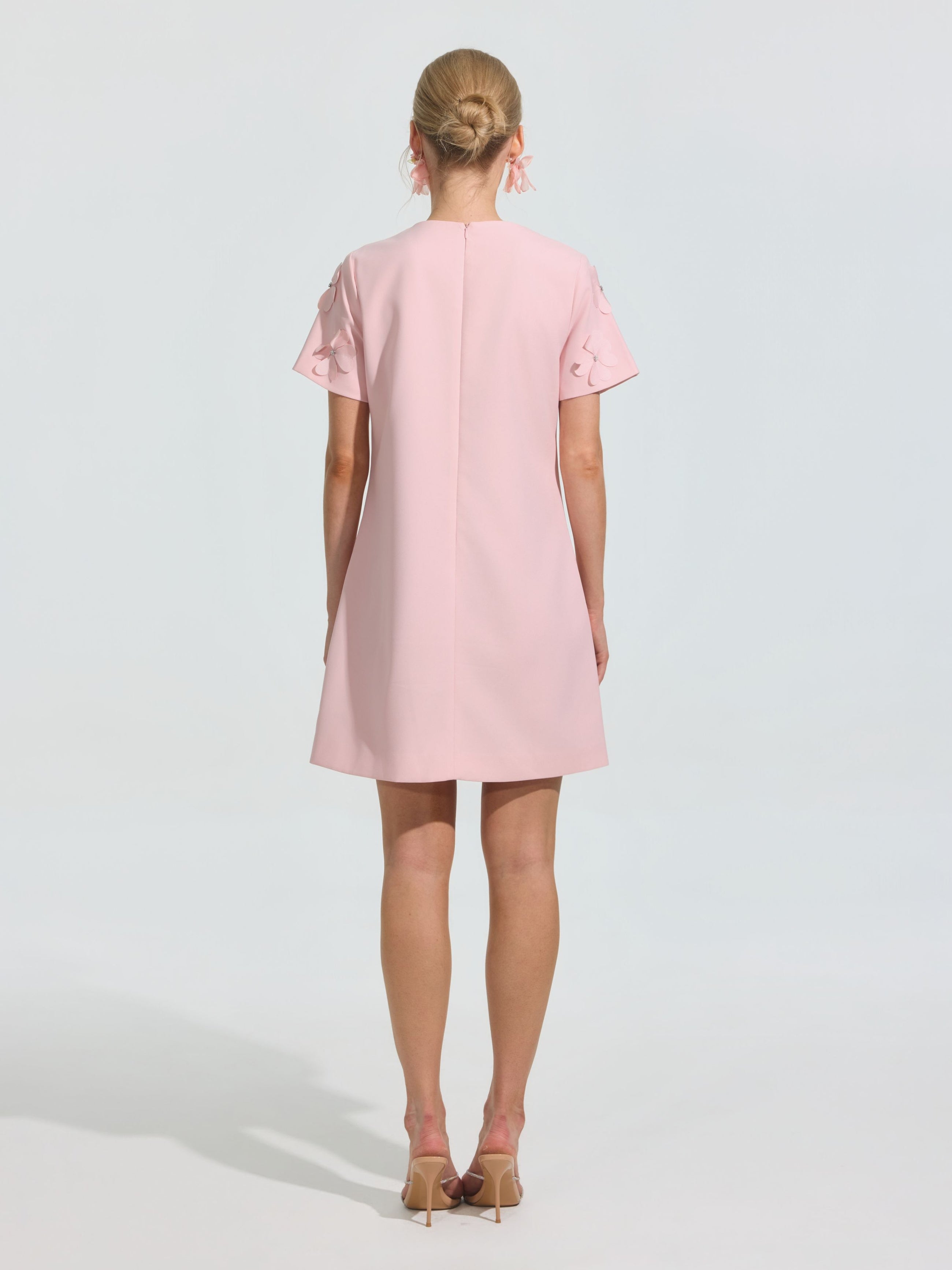 A woman faces away, wearing the Social Queen Beaded Floral Appliqué Dress—a short, light pink shift with short sleeves—paired with beige heels against a plain light background.
