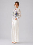 A woman stands against a plain light gray background wearing Social Queen’s Casual Wide-Leg Pants, paired with white heeled sandals and a sheer floral-embroidered jacket.