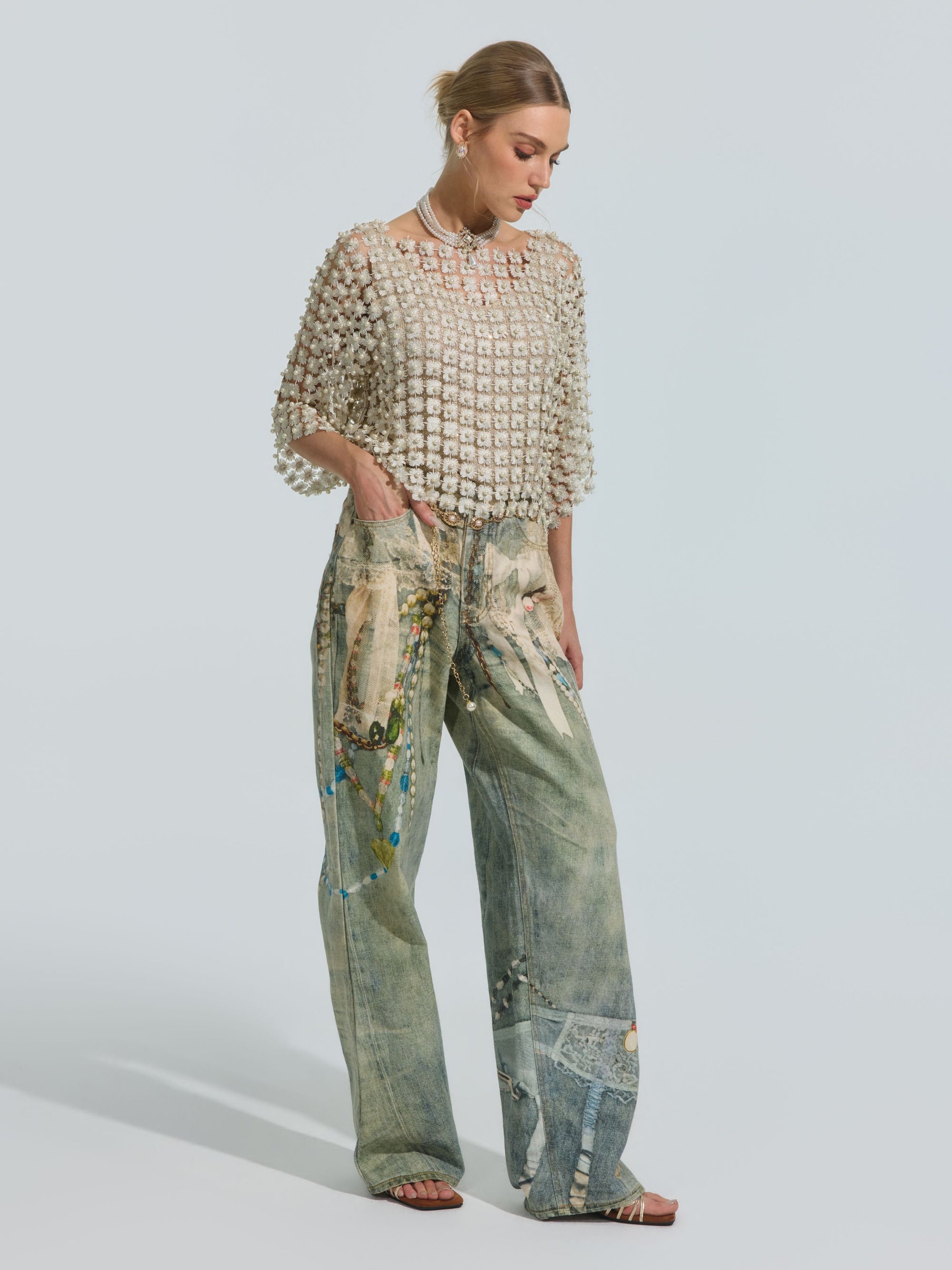 A woman stands in profile on a plain background wearing the Social Queen Pearl Daisy Cutwork Top (camisole not included), styled with loose, wide-legged patchwork and painted jeans, a necklace, and sandals.