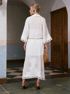 A woman stands indoors with her back to the camera, wearing the Social Queen Floral Cutwork Embroidered Linen Dress— a sheer white maxi dress featuring intricate floral cutwork embroidery. She holds a woven bag and pairs the look with heeled sandals.