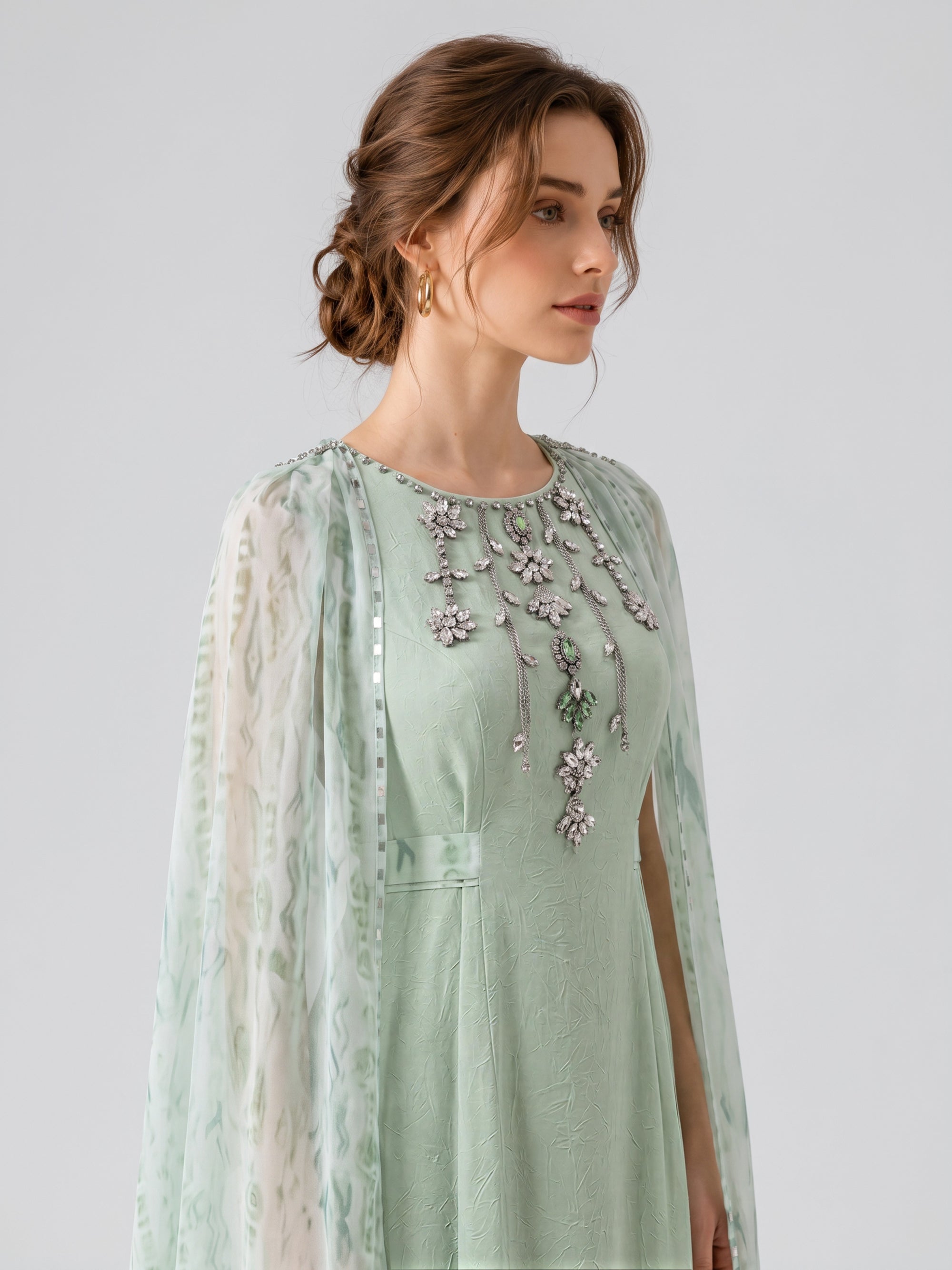 A woman with brown hair styled in an updo wears the Social Queen Beaded Printed Cape Chiffon Dress, a light green maxi dress adorned with jeweled floral embellishments and sheer patterned sleeves, finished with a delicate printed cape overlay. She poses against a plain light background.