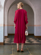 A woman stands indoors with her back to the camera, wearing the Social Queen 3D Floral Pearl Mulberry Silk Dress in deep pink. She carries a small decorative handbag, has blonde hair styled in a low bun, and wears heels.