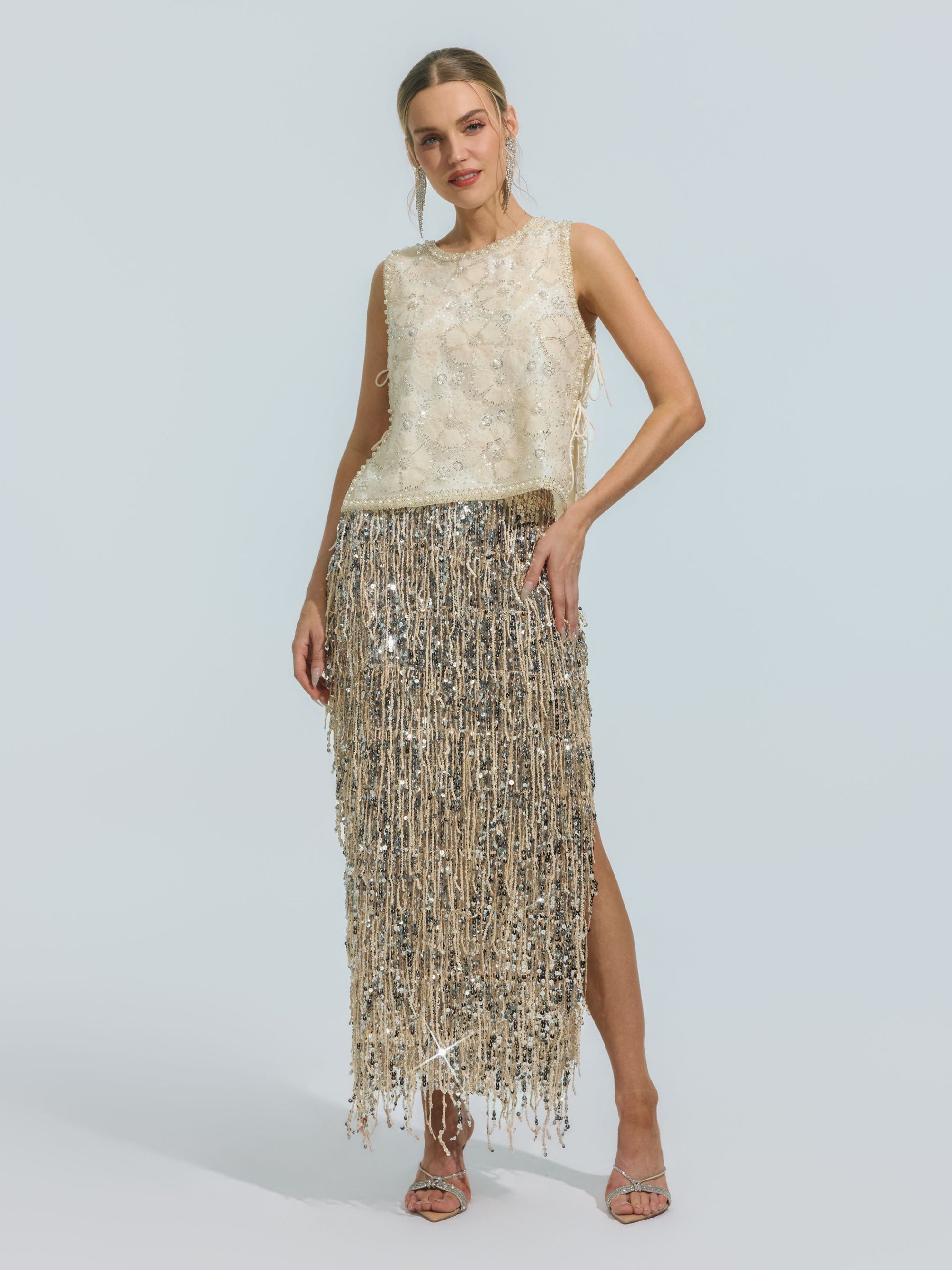 A woman stands in heels against a plain light background, wearing the Social Queen Floral Beaded Mesh Sleeveless Top—an ivory, lightweight mesh design with floral embellishments and delicate beaded details—paired with a metallic silver fringe skirt.