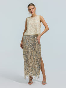 A woman stands in heels against a plain light background, wearing the Social Queen Floral Beaded Mesh Sleeveless Top—an ivory, lightweight mesh design with floral embellishments and delicate beaded details—paired with a metallic silver fringe skirt.