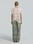 A woman stands facing away, wearing the Social Queen Pearl Rose Guipure Lace Top paired with loose-fitting, faded jeans adorned with decorative beads, chains, and subtle pearl details.