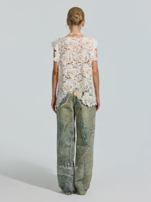 A woman stands facing away, wearing the Social Queen Pearl Rose Guipure Lace Top paired with loose-fitting, faded jeans adorned with decorative beads, chains, and subtle pearl details.