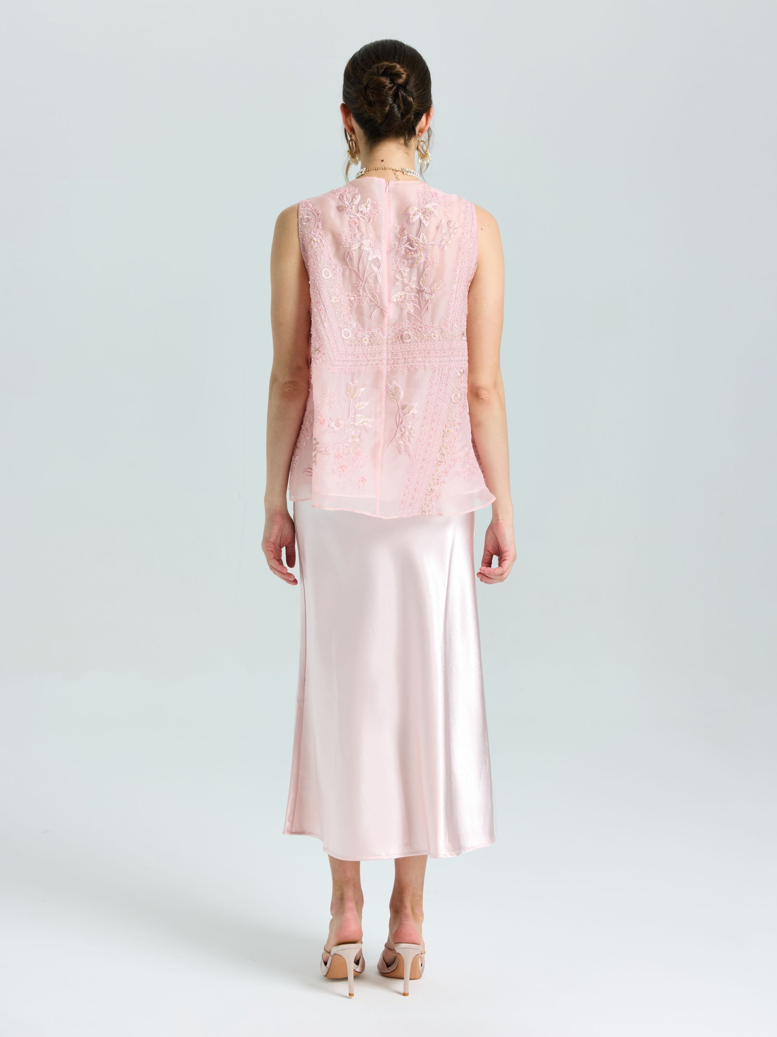 A woman stands facing away, wearing the Social Queen Floral Bird Embroidered Silk Top in light pink with lace details, paired with a satin midi skirt and nude high heels.