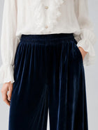 A person wearing a white ruffled blouse and Social Queen Silk Velvet Wide-Leg Pants in dark blue, with hands in pockets, shown from shoulders to thighs.