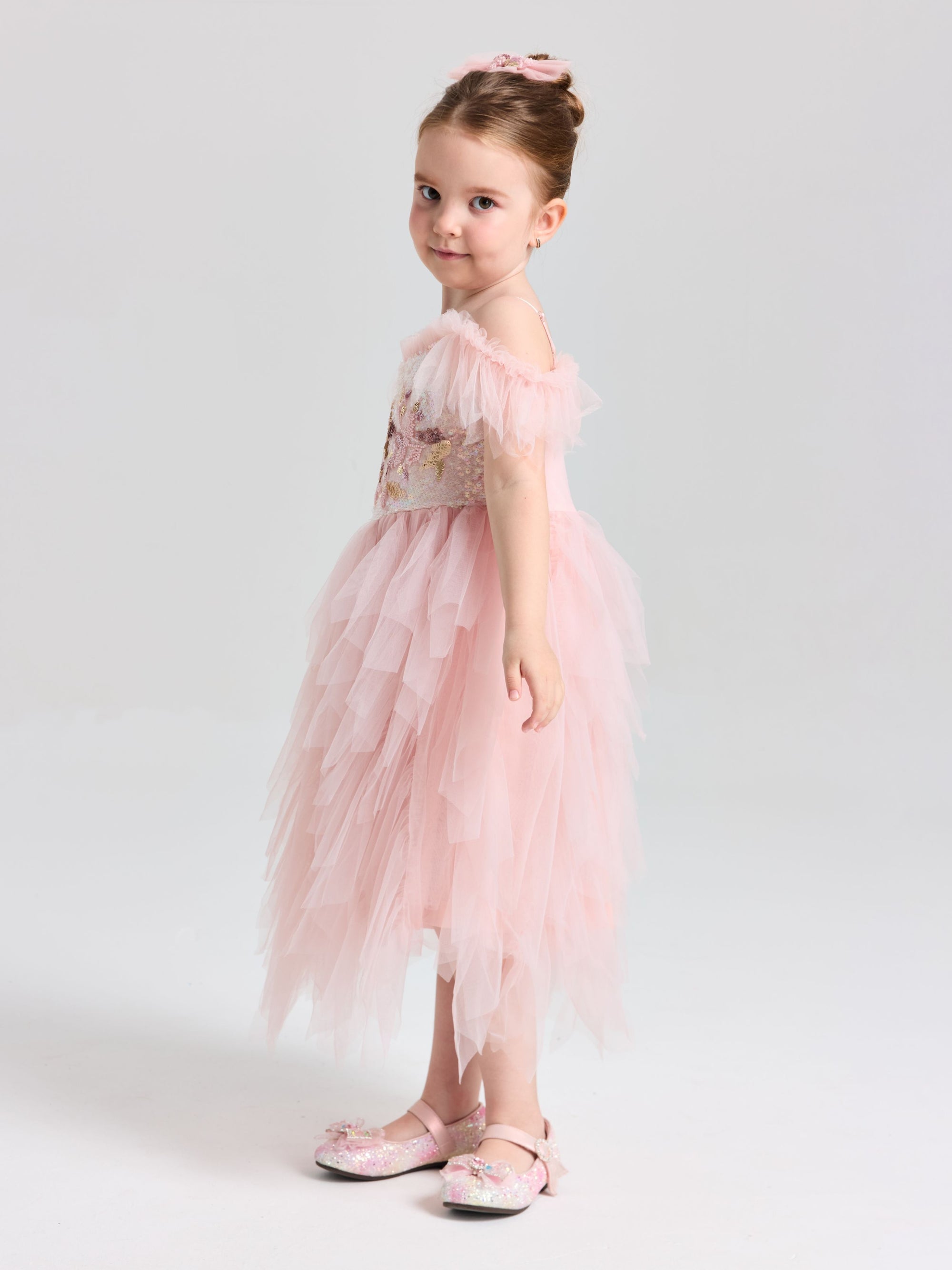A young girl poses against a light background in the Social Queen Girls Floral Sequin Tulle Dress, featuring an off-the-shoulder pink design with matching shoes.