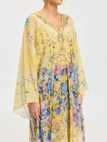 A woman wearing the Social Queen Floral Print Silk Caftan Dress—yellow, long-sleeved, V-neck and perfect for resort wear—stands against a plain background.