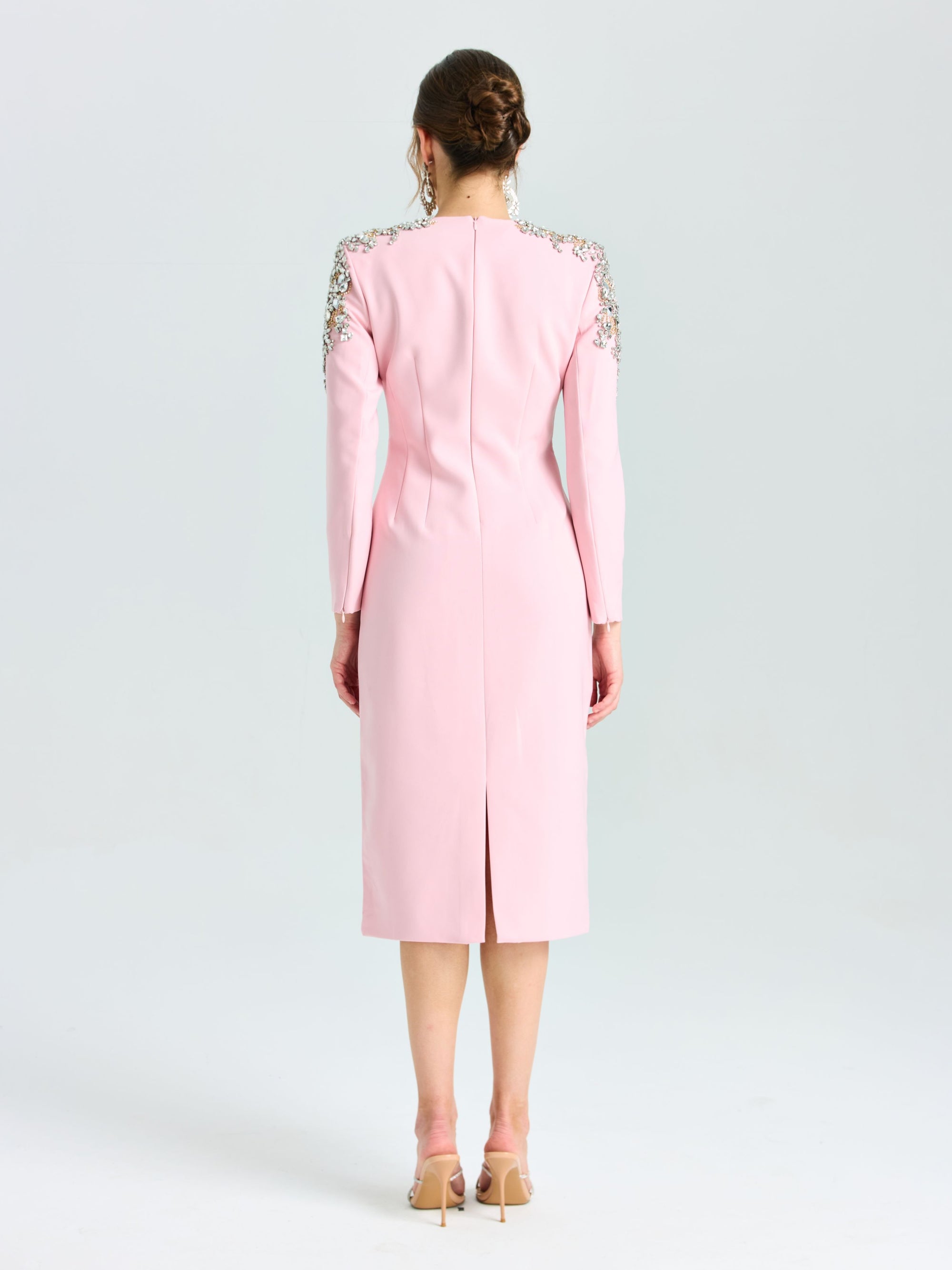 A woman stands facing away, wearing the Social Queen Crystal Beaded Detail Midi Dress in light pink crepe with long sleeves and crystal-embellished shoulders, styled with nude heels and a low bun—ideal for elegant evening occasions.