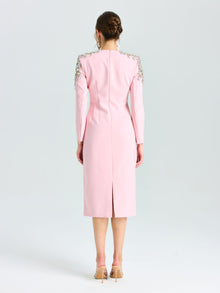 A woman stands facing away, wearing the Social Queen Crystal Beaded Detail Midi Dress in light pink crepe with long sleeves and crystal-embellished shoulders, styled with nude heels and a low bun—ideal for elegant evening occasions.