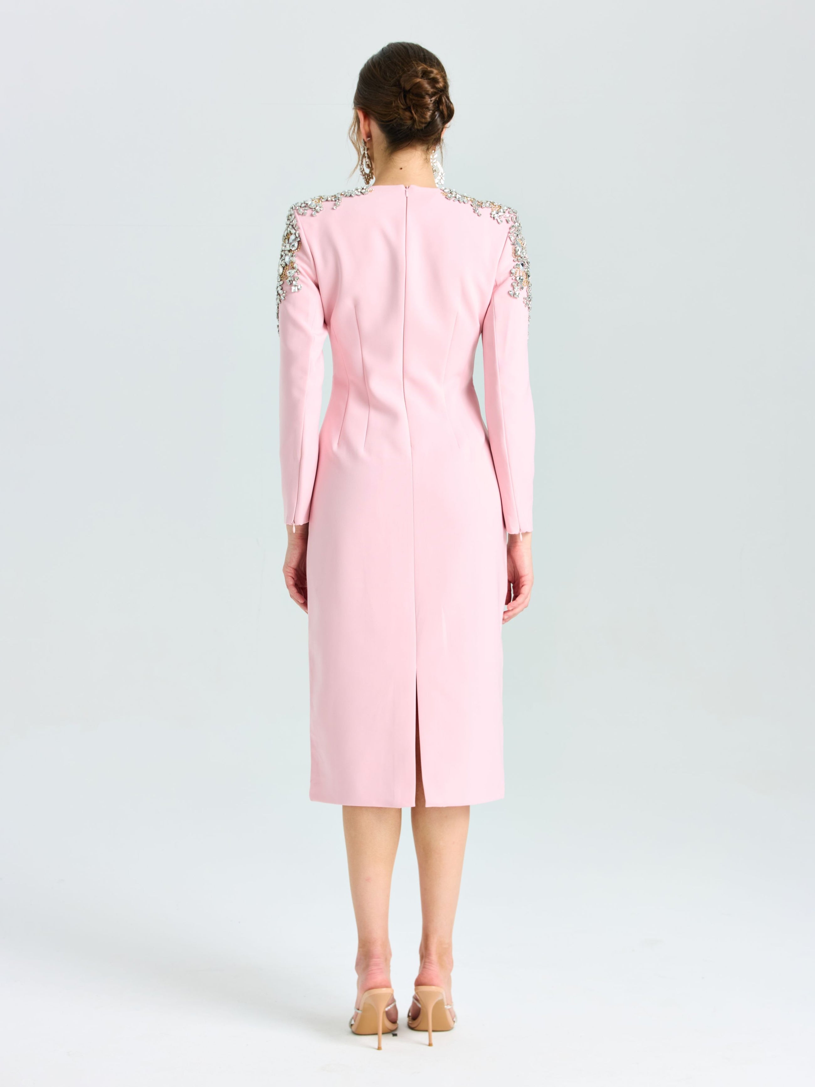 A woman stands facing away, wearing the Social Queen Crystal Beaded Detail Midi Dress in light pink crepe with long sleeves and crystal-embellished shoulders, styled with nude heels and a low bun—ideal for elegant evening occasions.
