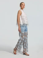 A woman stands in profile against a plain background, wearing the Social Queen 3D Floral Lace Appliqué Silk Top, paired with sheer lace wide-leg pants layered over light blue jeans and open-toed sandals.