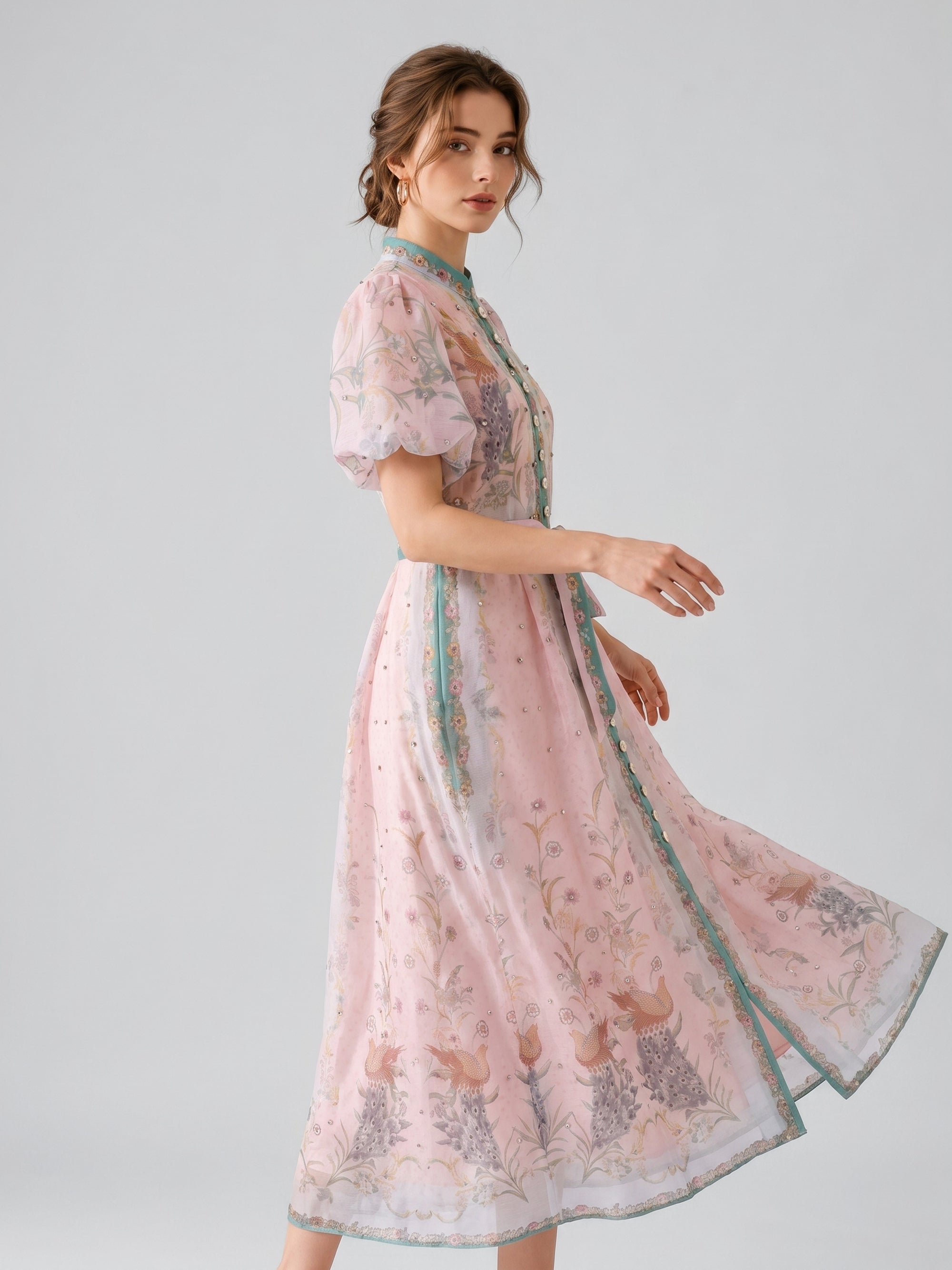 A woman wearing the Social Queen Peacock Botanical Print Dress, which features a pink base with floral embroidery, green trim, puff sleeves, and an elegant A-line silhouette with a coordinating belt, stands against a plain light gray background.