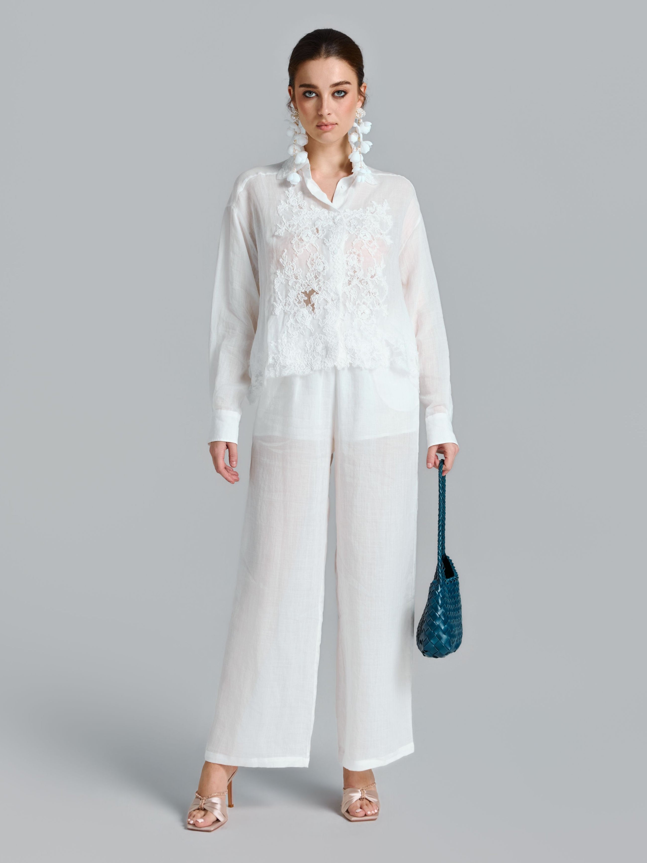 A woman stands against a gray background wearing the Social Queen 3D Lace Appliqué Shirt and wide-leg pants, holding a blue textured handbag, accessorized with statement earrings and heeled sandals.