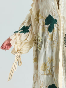 A person wears the Social Queen Floral Embroidered Linen Jacket (Cami Included), featuring cream linen, green floral embroidery, gold thread details, and a ribbon-tied cuff. Only the arm and torso are shown.