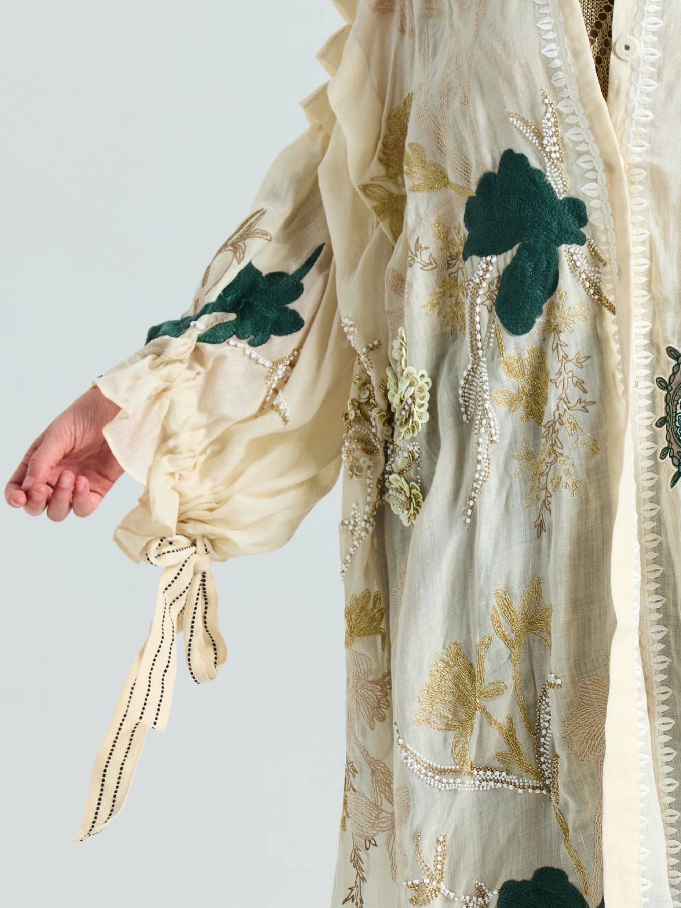 A person wears the Social Queen Floral Embroidered Linen Jacket (Cami Included), featuring cream linen, green floral embroidery, gold thread details, and a ribbon-tied cuff. Only the arm and torso are shown.
