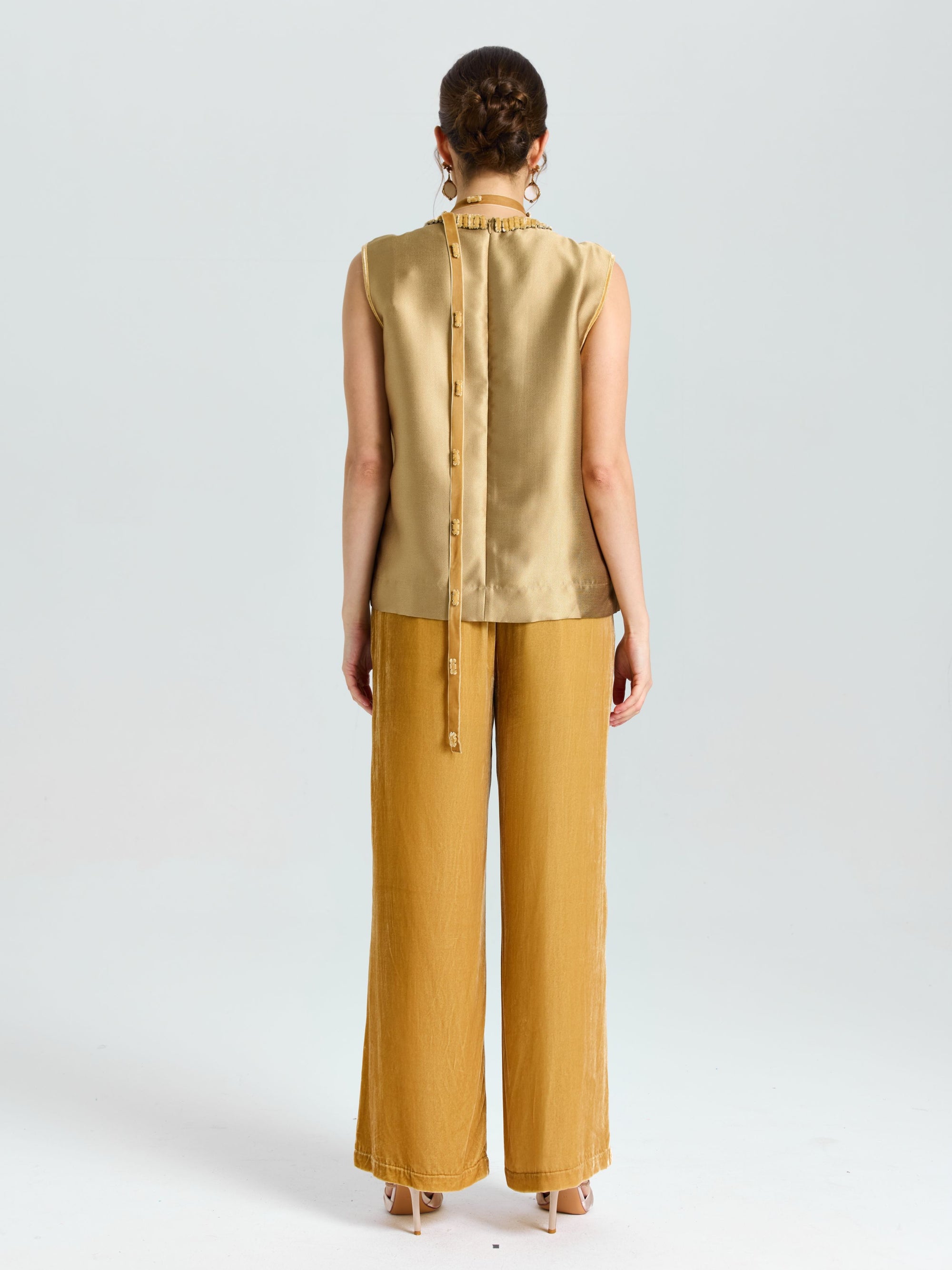 A person stands facing away, wearing a sleeveless gold top with button details down the back and Social Queen's Silk Velvet Wide-Leg Pants in mustard yellow, against a plain light background.