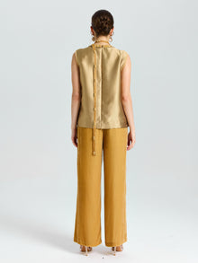 A person stands facing away, wearing a sleeveless gold top with button details down the back and Social Queen's Silk Velvet Wide-Leg Pants in mustard yellow, against a plain light background.