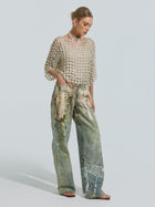 A woman stands in profile against a plain light background, wearing Social Queen’s Pearl Chain Printed Jeans—loose, wide-leg silhouette patchwork denim—paired with a sheer beaded top, necklace, and sandals.