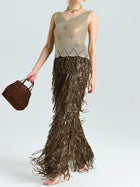 Model wears Social Queen’s Open-Knit Metallic Sleeveless Top with a long brown fringe skirt and holds a brown handbag, standing against a plain light background.